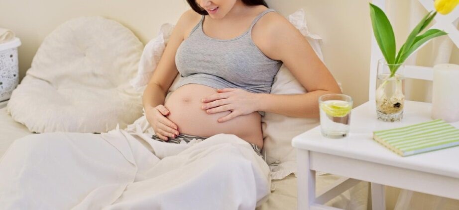 Pregnant woman sitting on bed holding her baby bump with a glass of water and notebook on the side table – IVF pregnancy precautions
