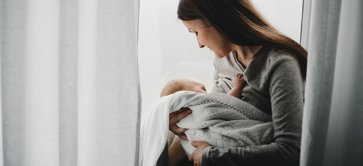Mother gently holding her newborn baby near a window, symbolizing postpartum care and emotional bonding at Rainbow Children’s Hospital.